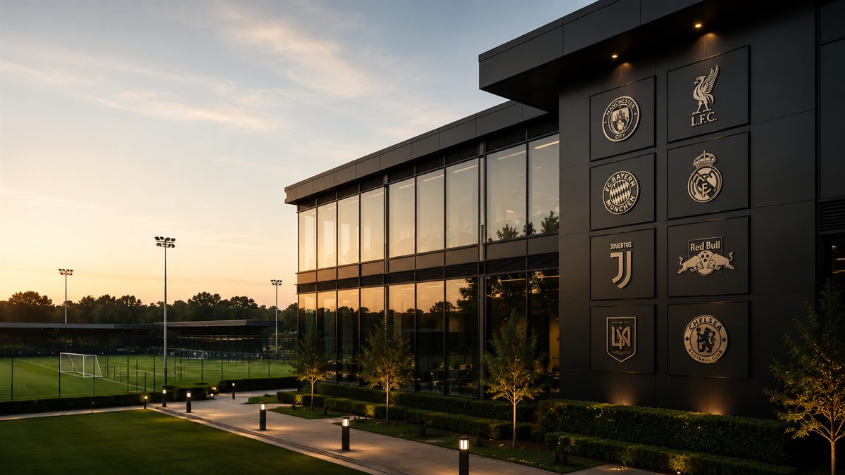 A modern football headquarters with club crests on its facade, overlooking training pitches at dusk